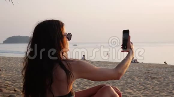 年轻的女人穿着泳衣和太阳镜带着手机坐在海滩上拍摄金色日落海的照片女孩视频的预览图