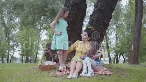 可爱优雅的老妇人坐在公园树下的毯子上和两个可爱的孙女交谈视频的预览图