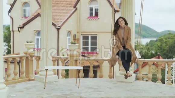 穿着米色外套的快乐年轻女子骑着秋千在欧洲城堡的院子里视频的预览图