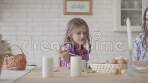 可爱的微笑的小女孩用一支小刷子在厨房里和妈妈坐在桌子旁画东方鸡蛋视频的预览图
