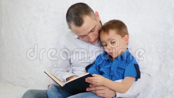 一位慈爱的父亲在家里的白色沙发上给他好奇的小儿子读一本有趣的书视频的预览图