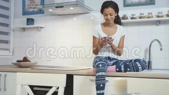 年轻的美国黑人妇女坐在厨房里在手机上写字视频的预览图