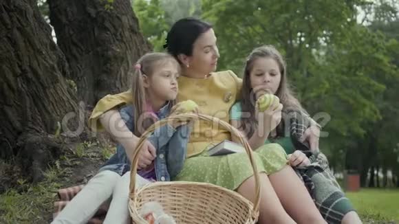 可爱的成熟女人和两个可爱的孙女坐在公园的树下吃苹果的女孩看着视频的预览图