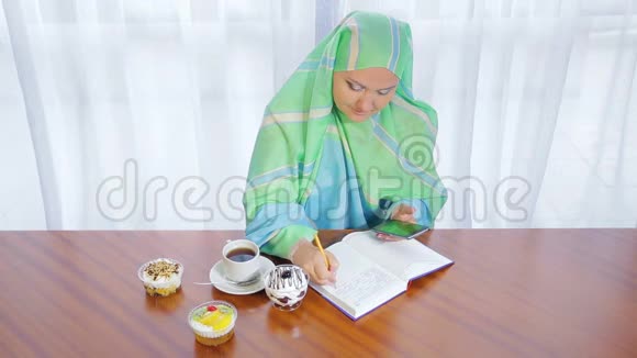 一个年轻的穆斯林妇女在咖啡馆里戴着一条浅色围巾一边用电话日记和喝茶一边远程工作视频的预览图