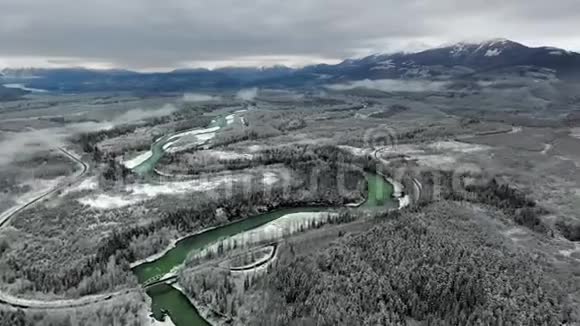 一架无人机正在一片茂密的松林一条河流和一条高速公路上旋转以观察冬天蓝雪覆盖的山脉视频的预览图