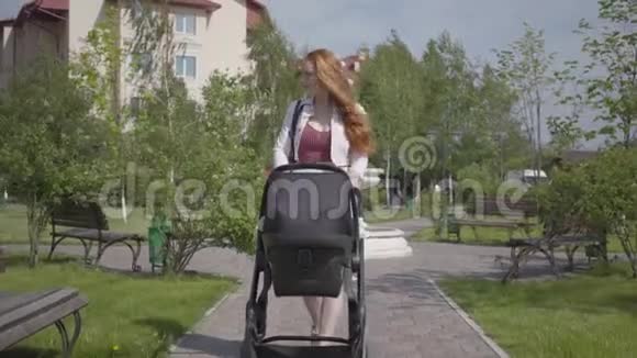 年轻快乐的红发妈妈在一个美好的春天带着婴儿车在街上散步微笑着视频的预览图