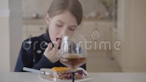 漂亮的金发女孩坐在厨房的桌子旁用她的手指吃蛋糕高高的玻璃杯和果汁站在附近视频的预览图