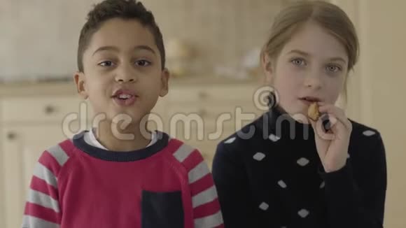 可爱的非裔美国男孩和金发白种人女孩蓝色的眼睛坐在厨房里吃饼干和饼干的女孩视频的预览图
