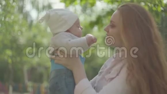 年轻美丽的红头发母亲抱着她可爱的孩子在她的怀里亲吻在春天阳光明媚的日子里和他说话视频的预览图