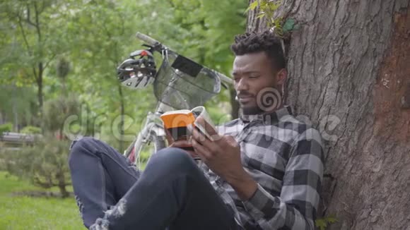 一位英俊自信的非洲裔美国人坐在公园的一棵老树下的自行车旁看书视频的预览图