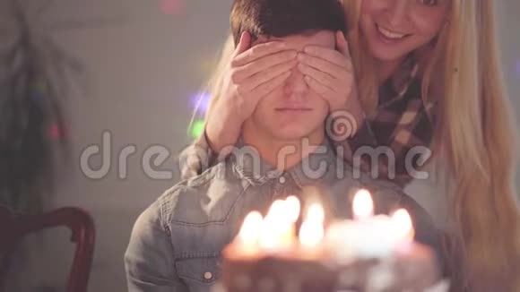 年轻的金发女孩遮住了帅哥的眼睛然后让他看到了有许多蜡烛的蛋糕他过生日视频的预览图