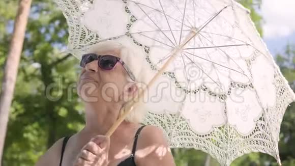 积极微笑的成熟女人戴着太阳镜站在公园的白色伞下环顾四周的画像视频的预览图