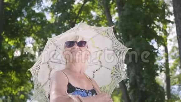 积极微笑的成熟女人戴着太阳镜站在公园的白色伞下环顾四周的画像视频的预览图