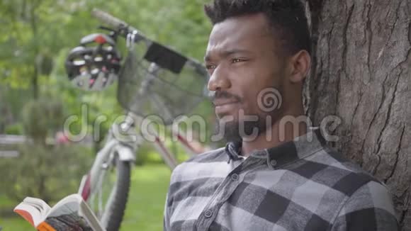 一位英俊的非洲裔美国人坐在公园的一棵老树下的自行车旁读着一本书视频的预览图