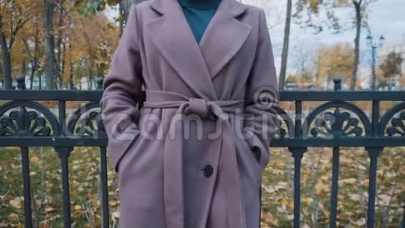 一个穿着淡紫色外套带着腰带的女孩正站在公园的篱笆前把外套合上视频的预览图