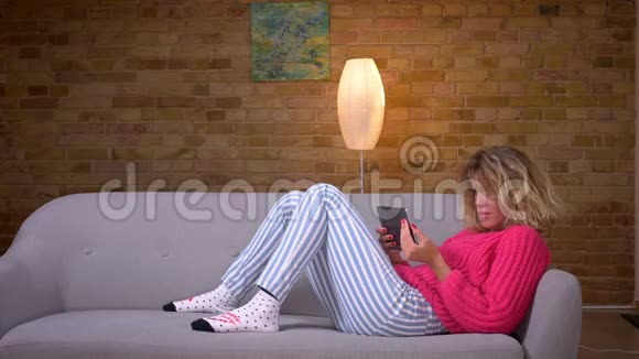 金发的家庭主妇穿着粉红色毛衣躺在沙发上在舒适的家庭氛围中在平板电脑上视频聊天视频的预览图