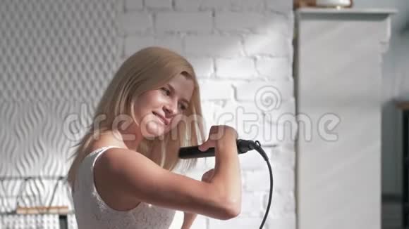 海尔多快乐迷人的女孩矫直头发与矫直机看相机在房间里视频的预览图