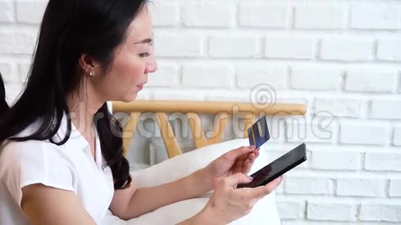 40多岁的亚洲中年妇女在舒适的白色房间里拿着信用卡和手机视频的预览图