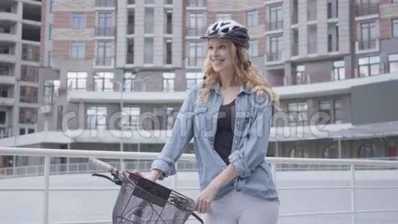 在城市建筑的背景下戴着自行车头盔的漂亮金发女人站在自行车上女士视频的预览图