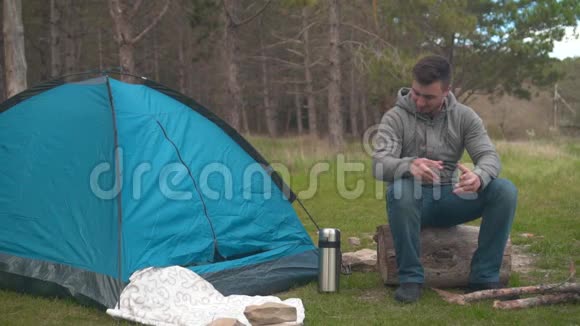 一个年轻人坐在森林里蓝色帐篷附近的一根大木头上从热水瓶里倒茶视频的预览图
