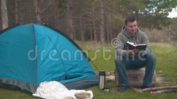 一个年轻人坐在一个大圆木附近的蓝色帐篷和Temos在森林里读一本书视频的预览图