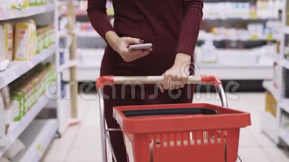 在超市的交易大厅里特写女士双手滚动红色手推车并使用电话视频的预览图