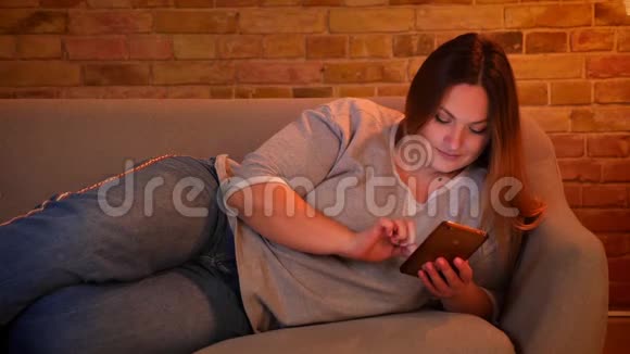 在舒适的家庭氛围中躺在沙发上愉快地在智能手机上聊天的轻松加码长发模特的肖像视频的预览图