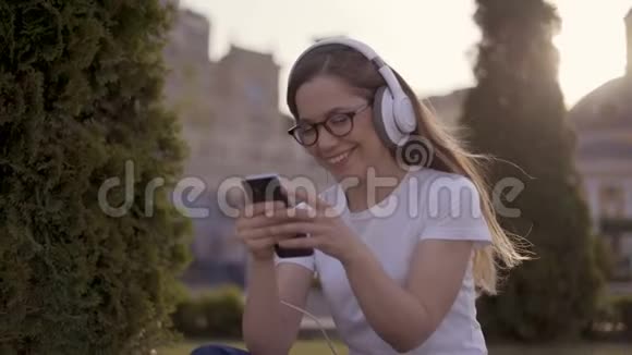 哇年轻惊讶的女孩戴着一个白色的大耳机坐在公园里女人打电话4K视频的预览图