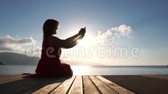 女人在日出时坐在浮舟上的智能手机上拍摄视频慢动作视频的预览图