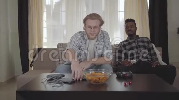 肖像英俊的胡子男坐在沙发上用手机说话而他的非裔美国人朋友正看着他视频的预览图