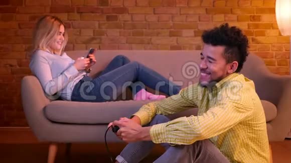 非洲男人坐在地板上玩电子游戏与操纵杆和高加索女孩工作与智能手机沙发在舒适的家视频的预览图