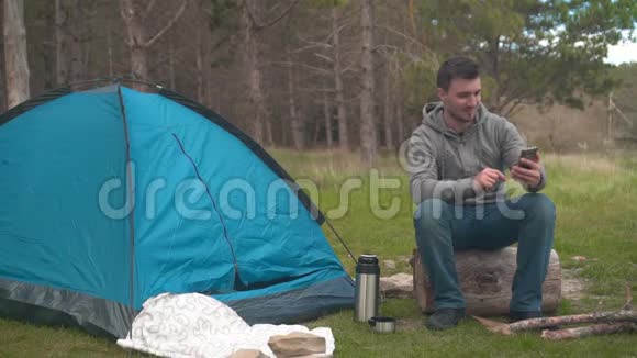 一个年轻人坐在蓝色帐篷附近的一根大木头上在电话上的视频链接上交谈视频的预览图