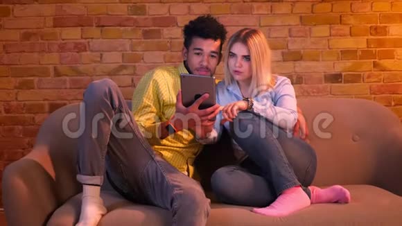 特写镜头拍摄的是非洲男人拿着平板电脑白种人金发女孩坐在家里的沙发上打字视频的预览图