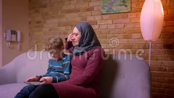 小男孩集中在平板电脑上玩游戏他的穆斯林母亲戴着头巾在家里观察他的活动视频的预览图