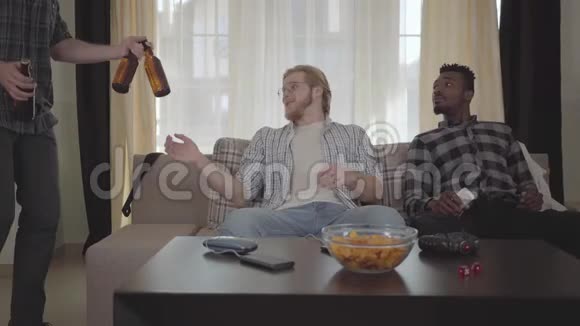 两个白种人和非裔美国人坐在沙发上第三个朋友带着啤酒瓶送给视频的预览图