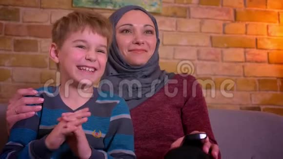 小男孩和他的穆斯林母亲戴着头巾的肖像在家里电视上笑着看喜剧视频的预览图