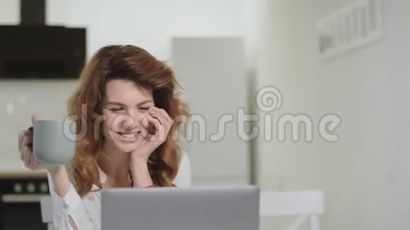 微笑的白人妇女在早上的厨房里的笔记本电脑上阅读快乐的新闻视频的预览图