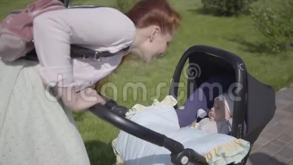漂亮的红发妈妈弯着腰在公园里的一辆电车上这位女士和她的孩子在户外享受阳光明媚的一天年轻母亲视频的预览图