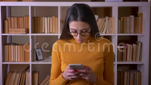 美丽的黑发老师戴着眼镜看着智能手机聚精会神地站在图书馆视频的预览图