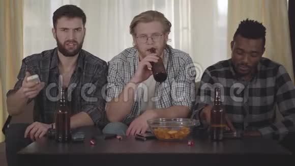 描绘了三个快乐的朋友坐在房子里看电视喝和薯片有一个视频电话视频的预览图