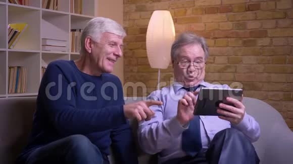 老年男性朋友坐在沙发上观看平板电脑并愉快地讨论的肖像视频的预览图