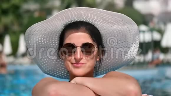 戴着太阳镜戴着白帽子在游泳池里享受日光浴的微笑女人视频的预览图