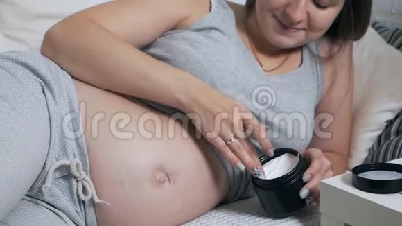 幸福的孕妇躺在家里的沙发上把奶油涂在肚子上视频的预览图