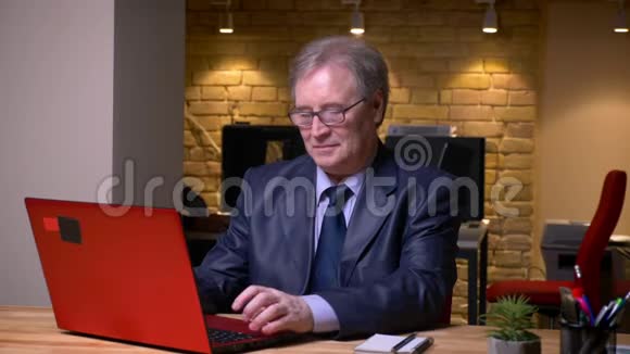 穿着正式服装在笔记本电脑上打字的老人的肖像在办公室里是快乐和积极的视频的预览图