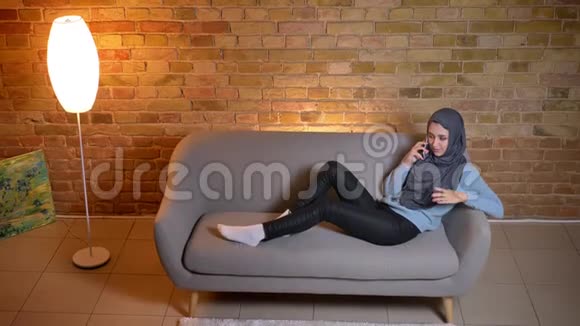 一位年轻迷人的穆斯林女性在舒适的家中躺在沙发上打着电话悠闲地休息视频的预览图