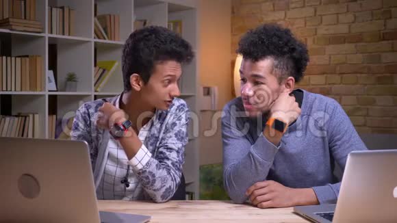 印度和非裔美国男学生使用笔记本电脑的特写镜头开心地坐着微笑着讨论视频的预览图