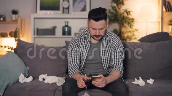 一个情绪低落的家伙一个人坐在家里用智能手机端着酒杯喝酒视频的预览图