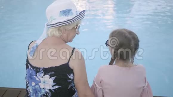 后景小滑稽女孩扎着辫子成熟的女人戴着太阳镜坐在池边休息视频的预览图