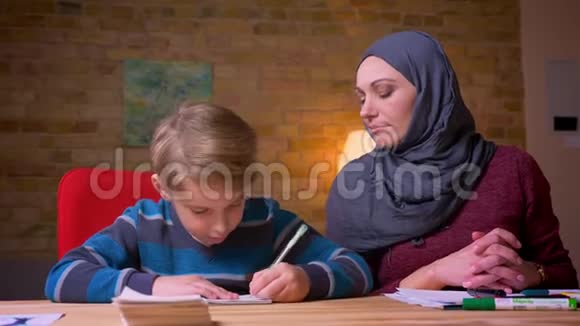 穆斯林母亲戴着头巾和她的小儿子在家里的桌子上做家庭作业视频的预览图