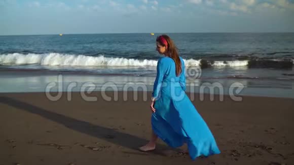 穿着漂亮蓝色连衣裙和太阳镜的女人在海滩上散步快乐地旋转着慢动作视频的预览图